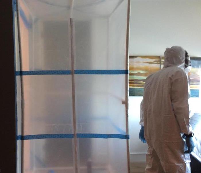 Protective plastic taped to ceilings and walls. Man in full white protective gear and blue gloves standing next to plastic.