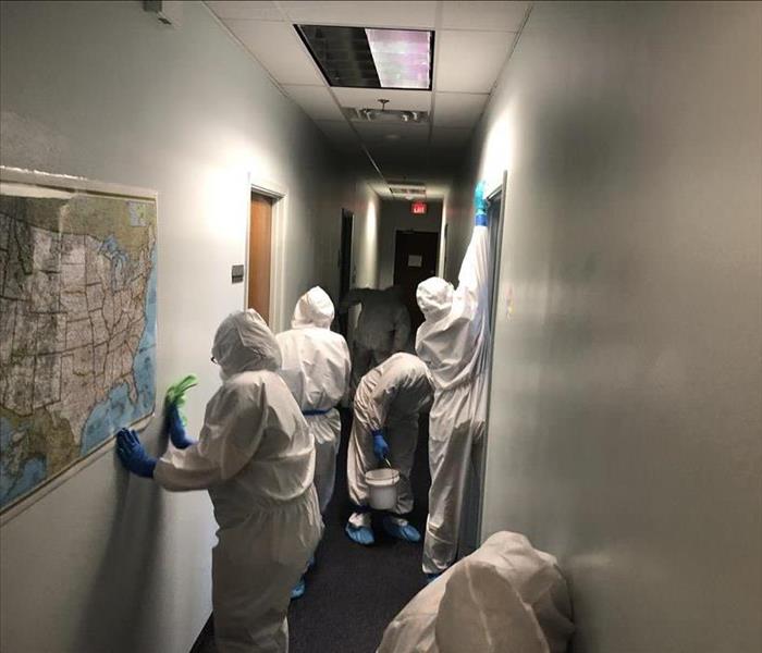 hallway in building, 6 people in full white protective body suits, wiping down walls.